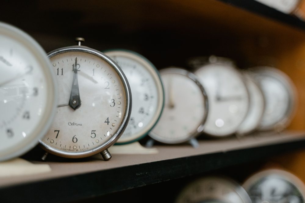 A row of vintage alarm clocks on a wooden shelf, the nearest in focus and the rest receding into blur