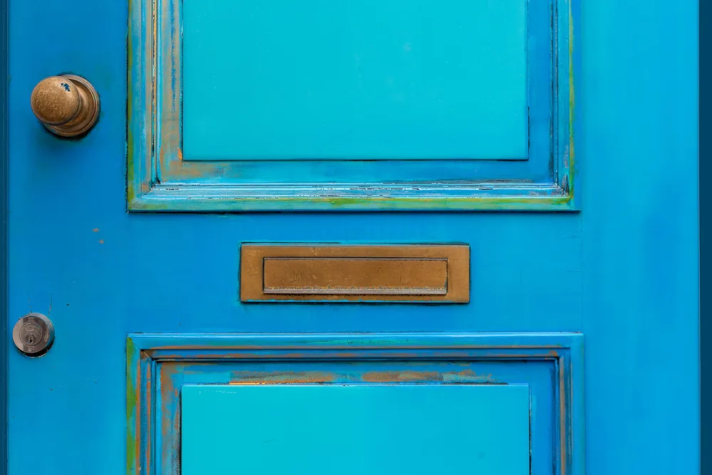 A brass letter slot in the centre of a weathered bright blue front door, with a brass knob and lock alongside